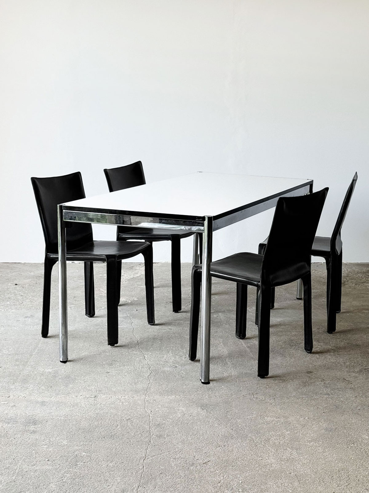 Stół USM Haller z chromowaną podstawą i białym laminowanym blatem, ustawiony z czterema czarnymi krzesłami Cassina.