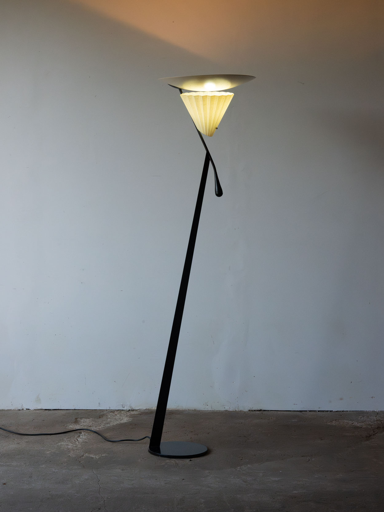 Włączona lampa podłogowa Mantide 315 Oluce z ciepłym światłem skierowanym ku górze i przez plisowany klosz.